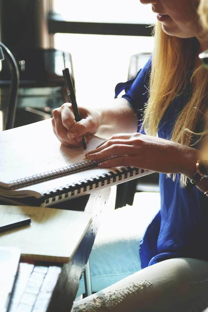 SIMONSAYS DIGITAL AGENCY Woman in a blue shirt writing in a spiral notebook at a wooden table in a bright cafe.