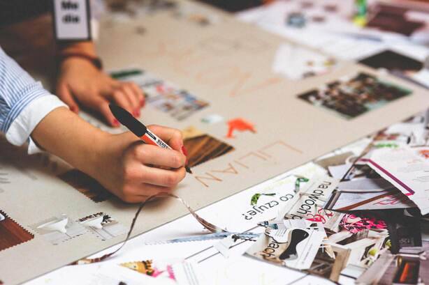 SIMONSAYS DIGITAL AGENCY A person writing on a large craft board surrounded by magazine clippings and materials for a creative project.