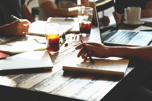 SIMONSAYS DIGITAL AGENCY Group collaboration at a coffee shop with laptops, notepads, and iced drinks on a wooden table.