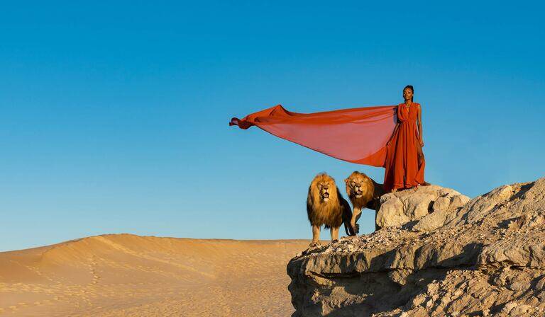 SIMONSAYS DIGITAL AGENCY A majestic woman in a flowing red dress stands on rocks with two lions against a clear blue sky.