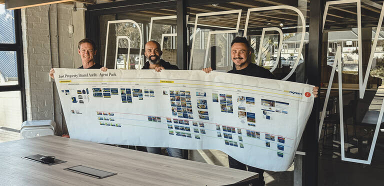 Just Property and SimonSAYS Creative Brand Agency team showcasing the full marketing campaign rollout plan, displayed on a large printed timeline, outside the agency’s modern glass-fronted office
